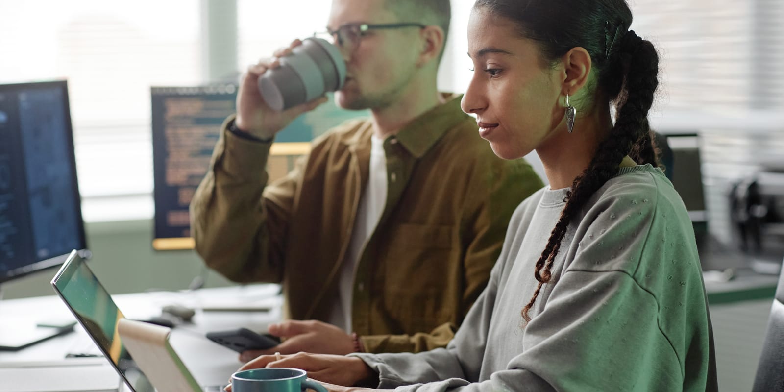 Two engineers at adjacent desks in an office with code on the monitors; one holds a coffee cup.