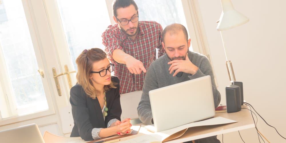 Three colleagues around a laptop at a sunlit desk, one pointing at the screen.