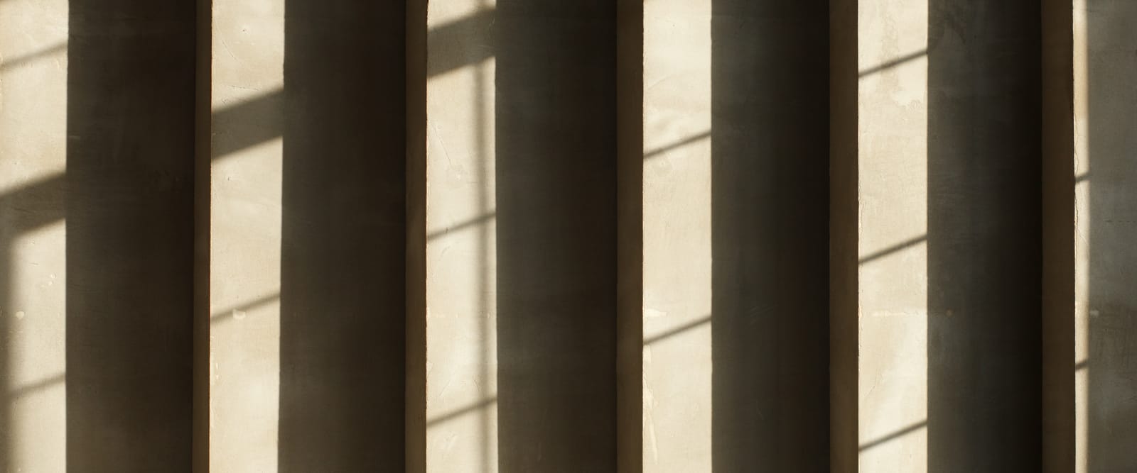 Sunlight through a row of windows casting evenly spaced bands of light and shadow on a concrete wall.